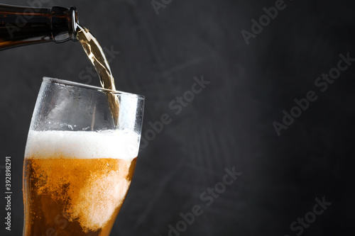 Pouring cold tasty beer from bottle into glass on black background, closeup. Space for text