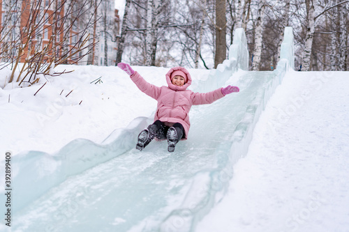 A young woman in a pink down jacket is riding in a city park on an ice slide without a sled
