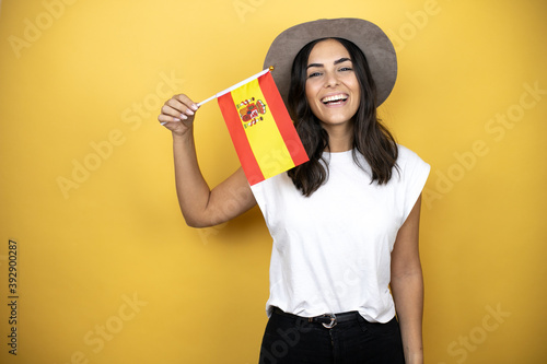 Beautiful woman wearing casual white t-shirt and a hat standing over yellow background holding the spanish flag