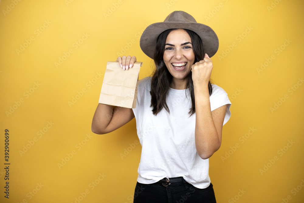 Beautiful woman wearing casual white t-shirt and a hat standing over yellow background holding a paper bag very happy and excited making winner gesture with raised arms Beautiful woman wearing casual white t-shirt and a hat standing over yellow background holding a paper bag very happy and excited making winner gesture with raised arms