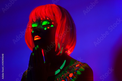 Close up profile portrait of a girl with artistic neon colorful makeup and orange wig, isolated violet blue background.
