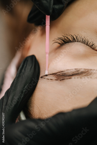 The girl s face with closed eyes. A master beautician in a medical gown  before microblading  corrects a sketch of an eyebrow on the girl s skin with a stick
