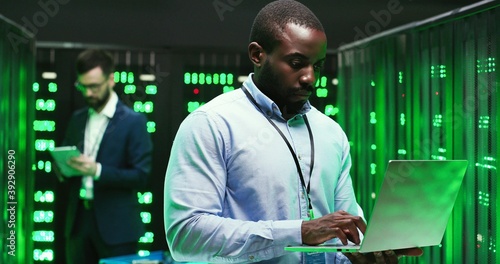 Portrait of handsome African American young man cryptograph standing in big data storage and working as typing on laptop computer. Male Caucasian analytic on background. Information security concept.