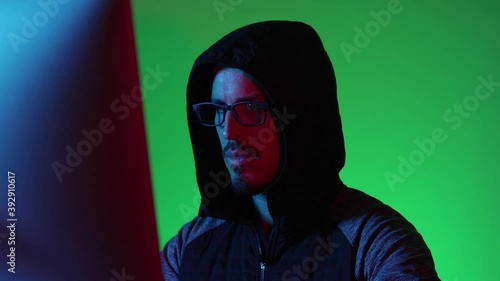 Hacker man sitting wearing hood and glasses, looking at the computer screen, typing and hacking. Working late night. Green background. Concept of dark internet, deep web. photo