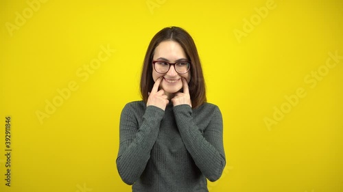 A young woman makes a cheerful smile on her face with her fingers on a yellow background. Woman with glasses and a green sweater. photo