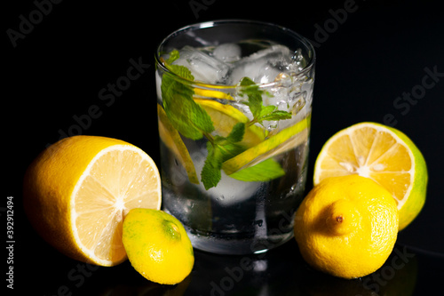 In the center of the screen are a glass of water, ice cubes, a sprig of mint and a slice of lemon. Cut lemons lie to the right and left. Closeup.
