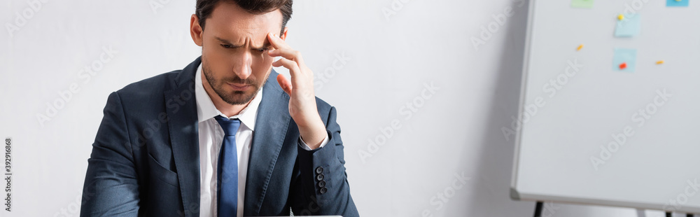Tired businessman with headache, holding hand near temple and looking away with blurred flipchart on background, banner Tired businessman with headache, holding hand near temple and looking away with blurred flipchart on background, banner