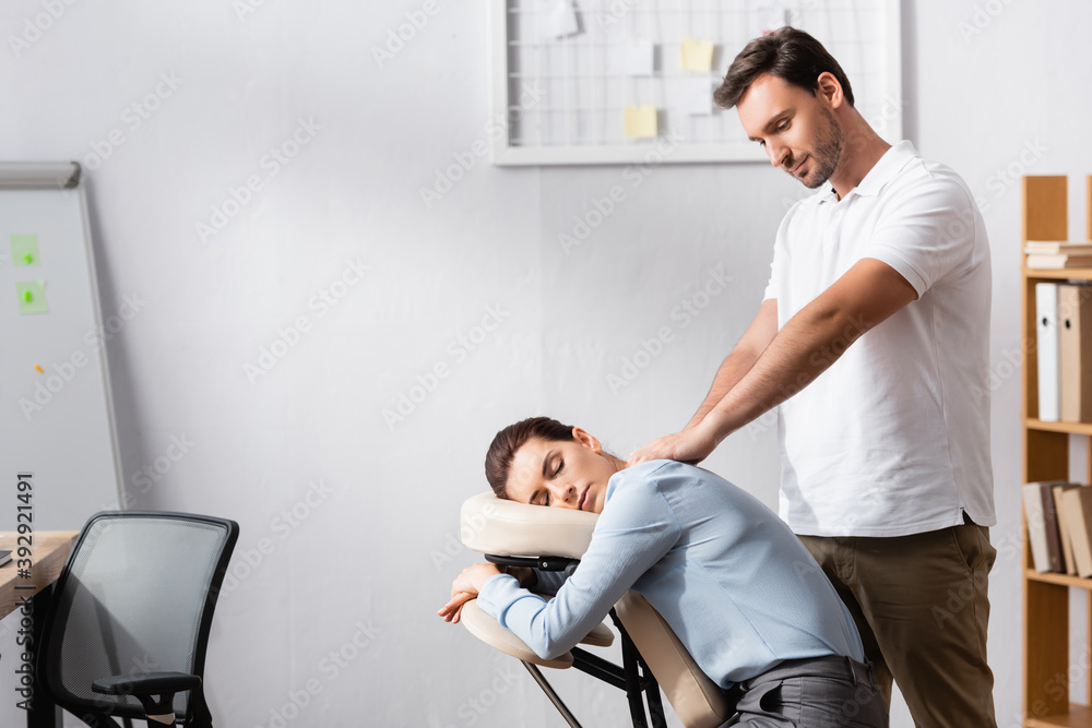 Fototapeta premium Smiling masseur with outstretched hands massaging shoulders of brunette woman sitting on massage chair in office