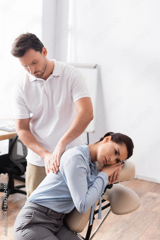 Obraz premium Massage therapist massaging back of businesswoman sitting on massage chair in office on blurred background