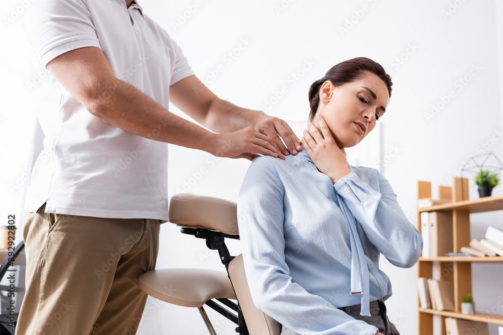 Fototapeta premium Massage therapist massaging painful shoulder of businesswoman siting on massage chair in office on blurred background