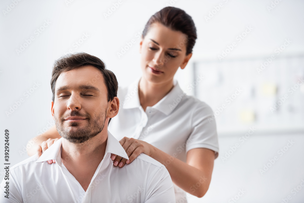 Masseuse massaging shoulders of smiling businessman with closed eyes on blurred background