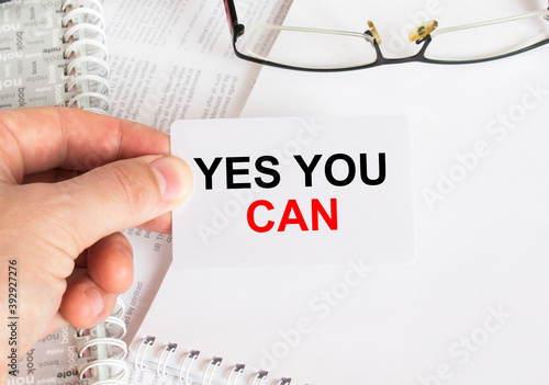 Man's hand holding a business card with text Yes You Can. Notepad with eyeglasses and text documents