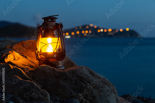 Frontal view of an old oil lantern sitting on a rock of the beach of Cala Regina and city lights bokeh on the background photo