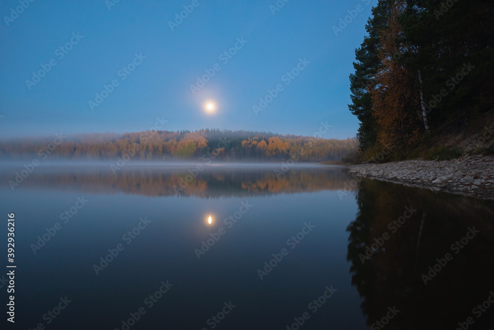 Obraz premium Scenic view of the river banks with trees covered in autumn colored leafs and mist during sunrise and full moon set