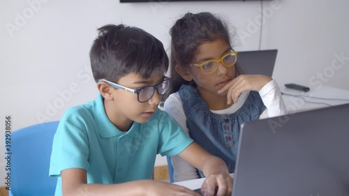 Couple of schoolchildren in glasses working on task together, using laptop in classroom, watching content and laughing. Static shot. Digital education concept