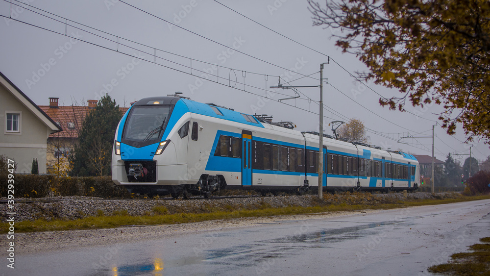 Modern white and blue passenger commuter train passing by in rain. Rainy commute with a modern train in cold and grey weather.