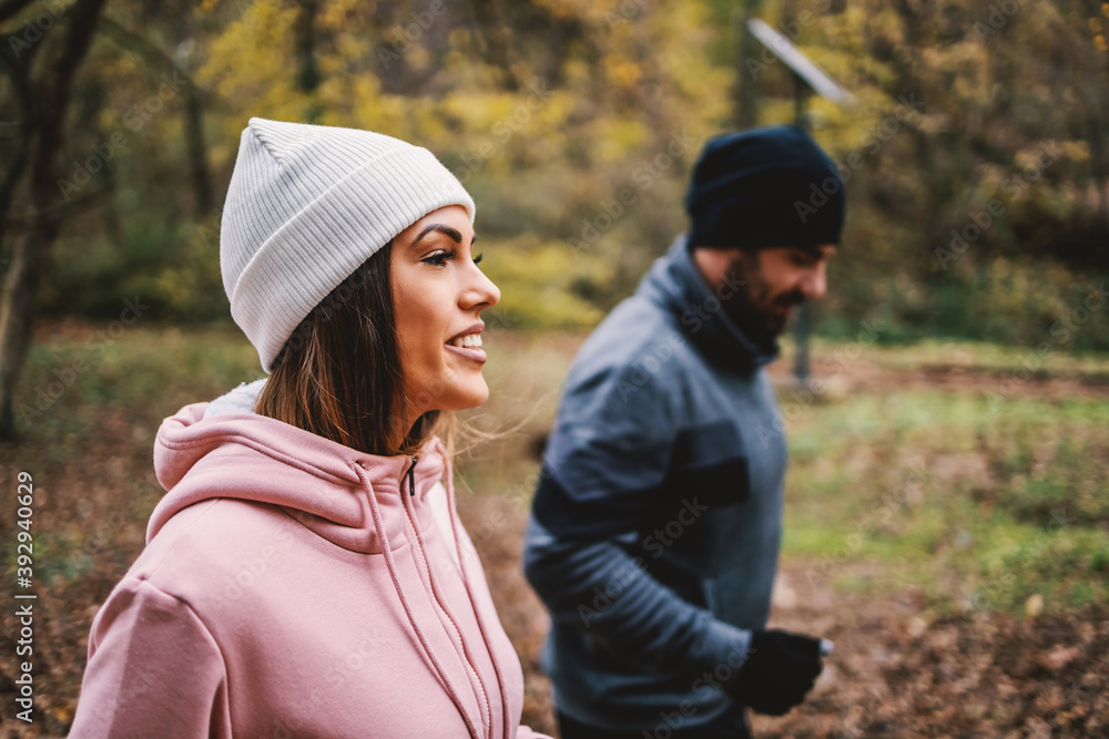 Fototapeta premium Sporty happy couple in warm outfit jogging in woods at autumn. Selective focus at woman.