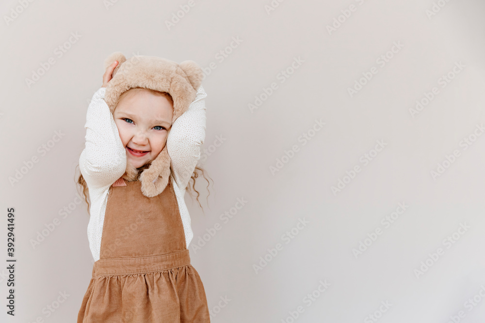 Little cute girl in a fur hat with an abalone on a beige background. A 3 year old child is holding his head.