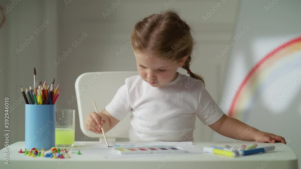 Lovely little girl using a brush and watercolors to paint on paper. Smart concentrated kid spending time, studying at home. Image of rainbow on background. Lovely little girl using a brush and watercolors to paint on paper. Smart concentrated kid spending time, studying at home. Image of rainbow on background.