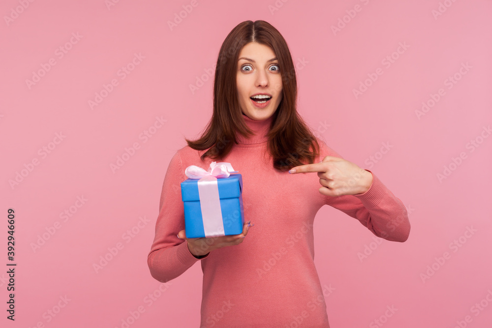 Excited surprised brunette woman in pink sweater pointing finger at blue giftbox in her hand looking at camera with wondered expression. Indoor studio shot isolated on pink background