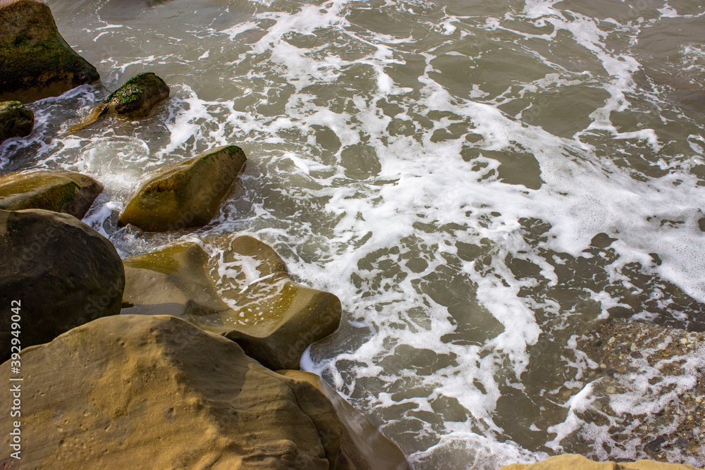 Obraz premium Stormy sea. Powerful big foam waves beat against big beautiful rocks, rocks, breakwaters. A scattering of spray. Cloudy weather with bright light.