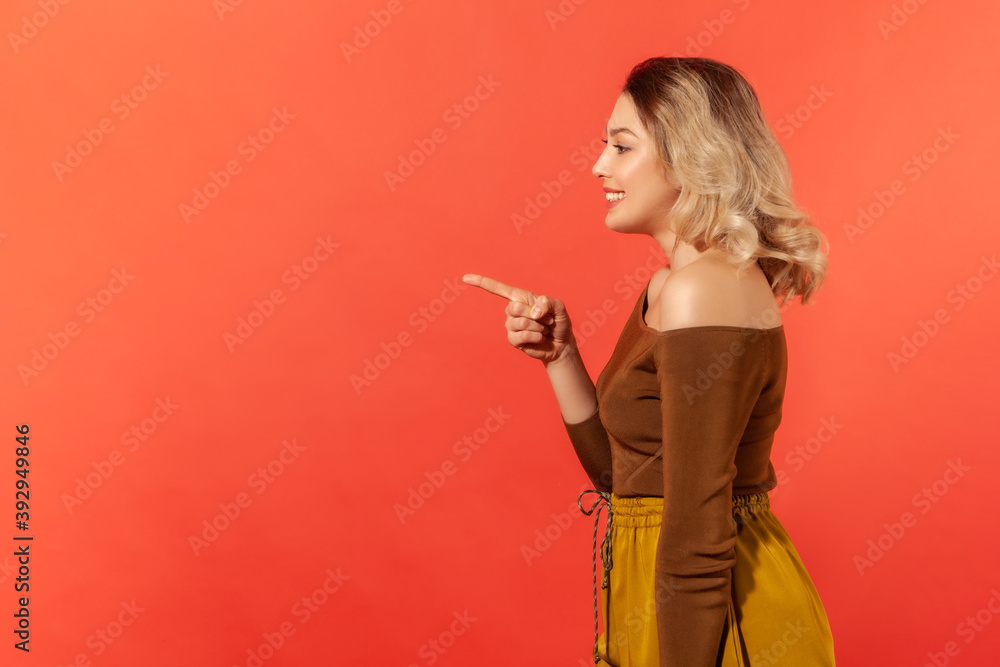 Side view portrait of beautiful toothy smiling woman with blonde hair in brown blouse pointing finger to the left, looking with astonishment. Indoor studio shot isolated on red background Side view portrait of beautiful toothy smiling woman with blonde hair in brown blouse pointing finger to the left, looking with astonishment. Indoor studio shot isolated on red background