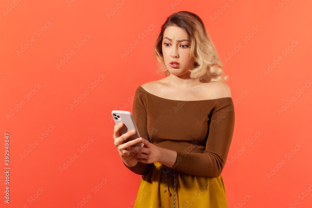 Upset frustrated woman looking at smartphone display in her hands, reading unpleasant message or email, dissatisfied with unfavorable tariffs. Indoor studio shot isolated on orange background Upset frustrated woman looking at smartphone display in her hands, reading unpleasant message or email, dissatisfied with unfavorable tariffs. Indoor studio shot isolated on orange background