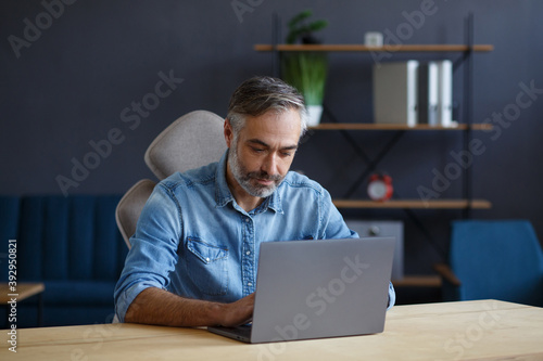 Grey-haired senior man working in home office with laptop. Business portrait of handsome manager sitting at workplace. Studying online, online courses. Business concept.