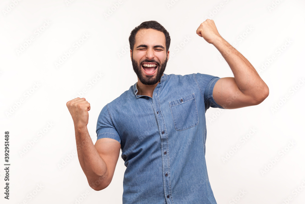 Fototapeta premium Euphoric thrilled man with beard screaming hurray raising hands up, celebrating his triumph and victory, extremely satisfied with success. Indoor studio shot isolated on white background