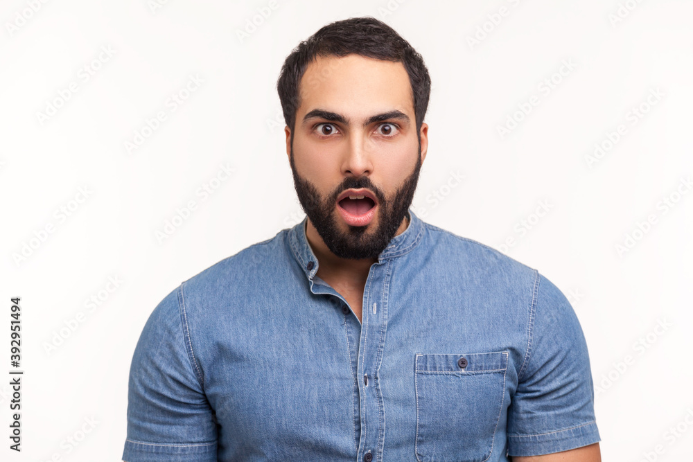 Fototapeta premium Surprised wondered man with beard in blue shirt looking at camera with big eyes and open mouth, shocked with unexpected news. Indoor studio shot isolated on white background