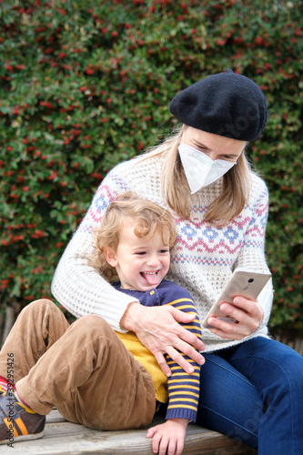 Mother with child on park bench, using mobile phone, Woman with protective mask, coronavirus prevention