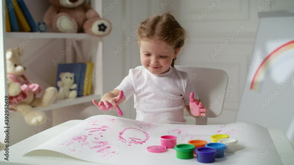 Lovely happy little girl preschooler uses fingers covered in pink paint to create cute abstract images on big sheet of paper. Kid spending time at home with pleasure. Lovely happy little girl preschooler uses fingers covered in pink paint to create cute abstract images on big sheet of paper. Kid spending time at home with pleasure.