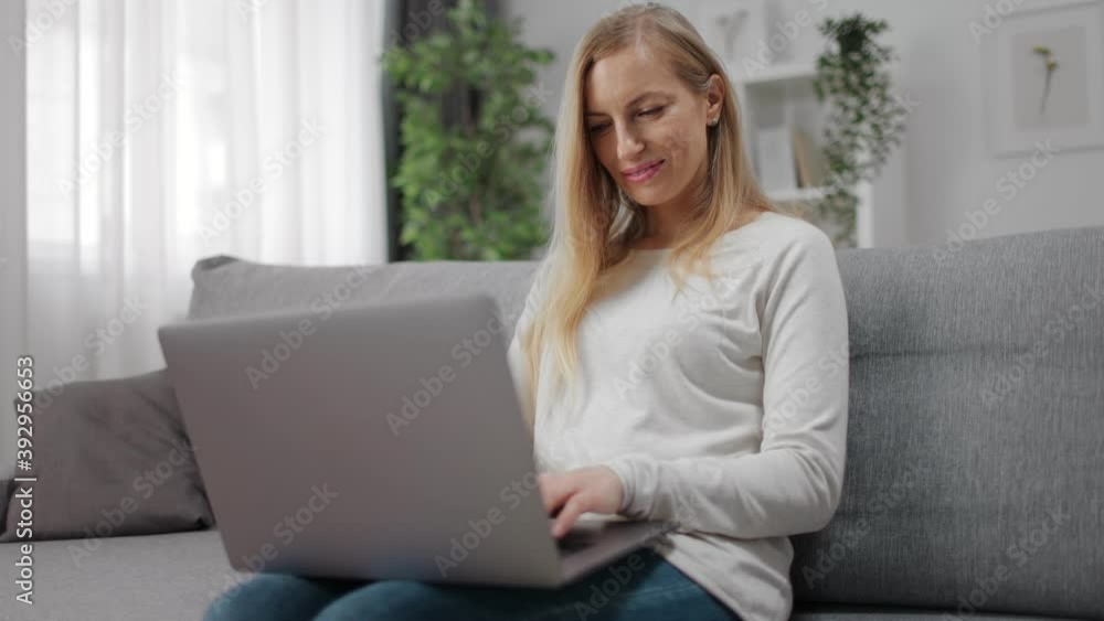 Smiling woman with blond hair sitting on cozy sofa and typing on portable laptop. Happy female using wireless gadget while staying at home. Smiling woman with blond hair sitting on cozy sofa and typing on portable laptop. Happy female using wireless gadget while staying at home.