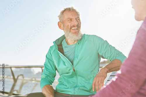 Cheerful sporty senior couple resting, sitting on the bench after workout outdoors. Mature man smiling to woman while discussing something