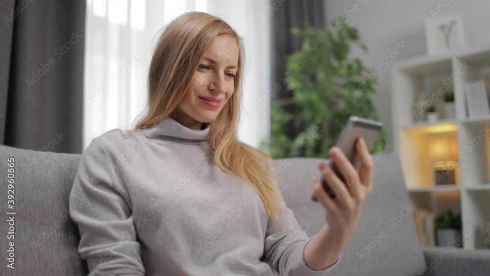 Happy woman with blond hair relaxing on grey couch and using smartphone. Smiling lady spending leisure time at home with modern gadget. Happy woman with blond hair relaxing on grey couch and using smartphone. Smiling lady spending leisure time at home with modern gadget.