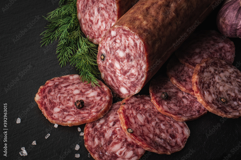 Sliced salami sausage on a board. Meat product and herbs on a dark back. A quick meal or snack. Dried sausage with black peppercorns. Sliced salami sausage on a board. Meat product and herbs on a dark back. A quick meal or snack. Dried sausage with black peppercorns.