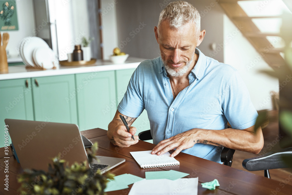 Focused middle aged caucasian man making some notes while sitting at table at home during remote working day Focused middle aged caucasian man making some notes while sitting at table at home during remote working day
