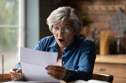 Close up overjoyed mature woman wearing glasses holding paper sheet, shocked surprised grey haired female with open mouth reading unexpected news in letter, money refund, sitting at desk