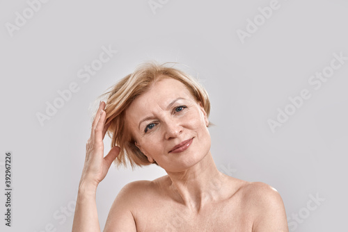 Portrait of beautiful middle aged woman looking at camera, adjusting her hair, posing isolated against grey background. Beauty, skincare concept