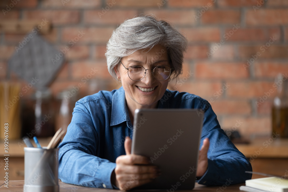 Obraz premium Close up smiling mature woman wearing glasses using computer tablet, chatting or shopping online, looking at screen, happy senior female having fun with device, browsing apps, making video call