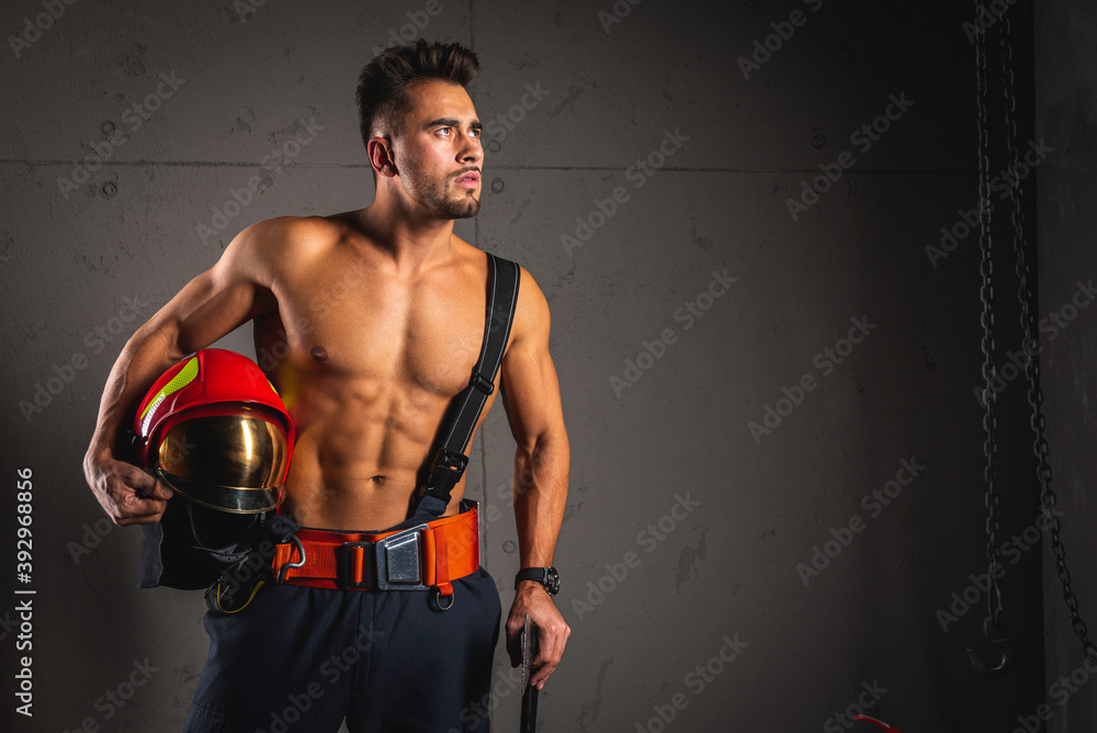 portrait of a muscular, handsome firefighter in uniform on a dark background holding a fire helmet, junk, looking away. low key, selective focus. The profession of firefighter