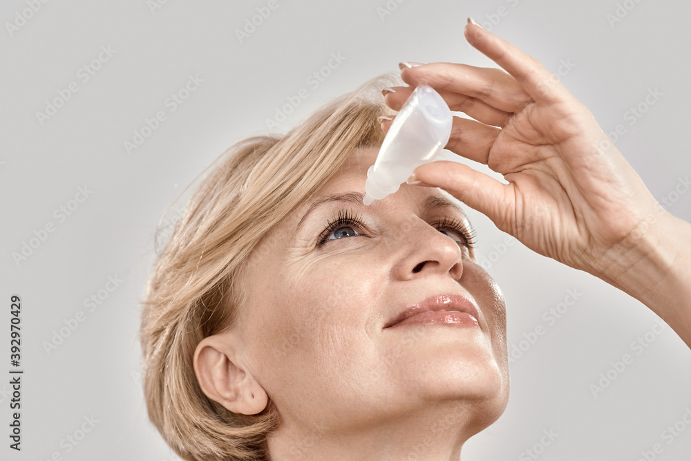 Close up portrait of attractive middle aged woman applying eye drops, standing isolated over grey background