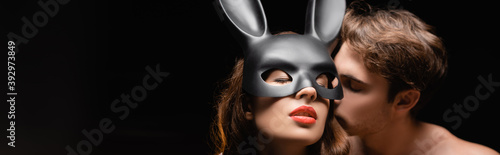 Shirtless man kissing sexy woman in rabbit mask with red lips isolated on black, banner photo