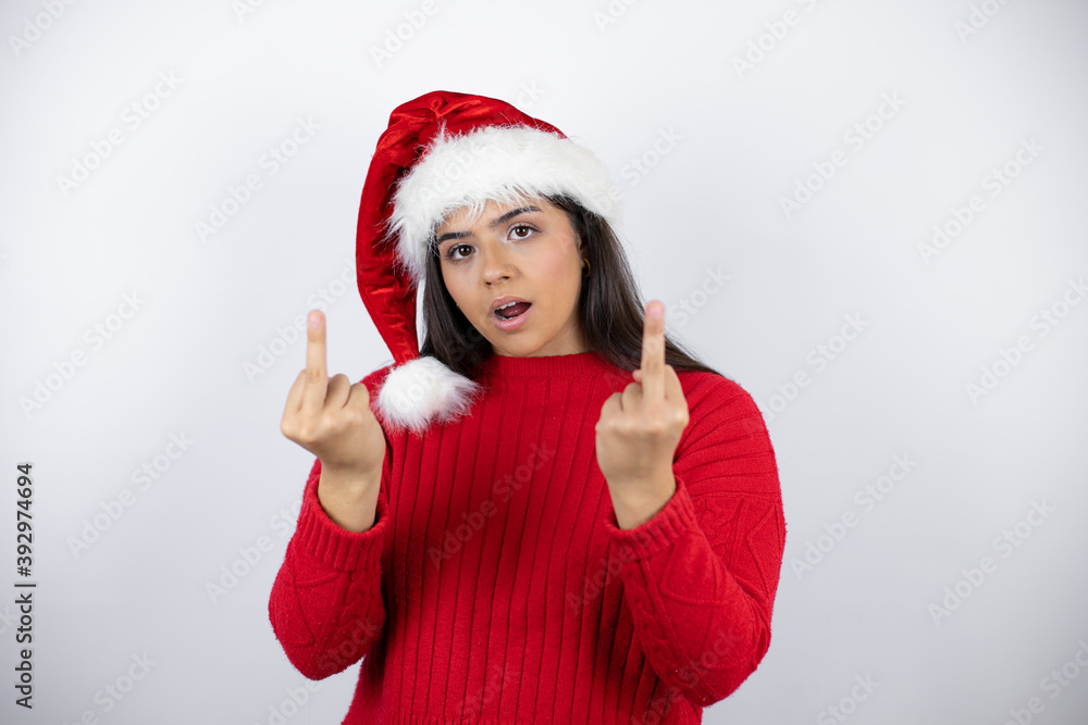 Young beautiful woman wearing a Santa hat over white background showing middle finger doing fuck you bad expression, provocation and rude attitude. screaming excited Young beautiful woman wearing a Santa hat over white background showing middle finger doing fuck you bad expression, provocation and rude attitude. screaming excited