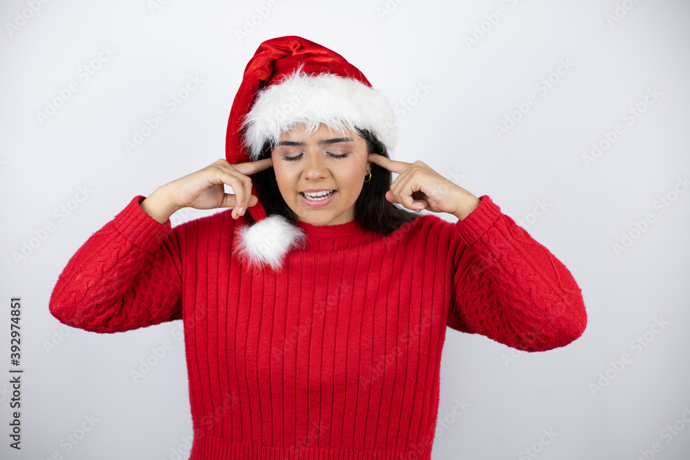 Young beautiful woman wearing a Santa hat over white background covering ears with fingers with annoyed expression for the noise of loud music. Deaf concept. Young beautiful woman wearing a Santa hat over white background covering ears with fingers with annoyed expression for the noise of loud music. Deaf concept.
