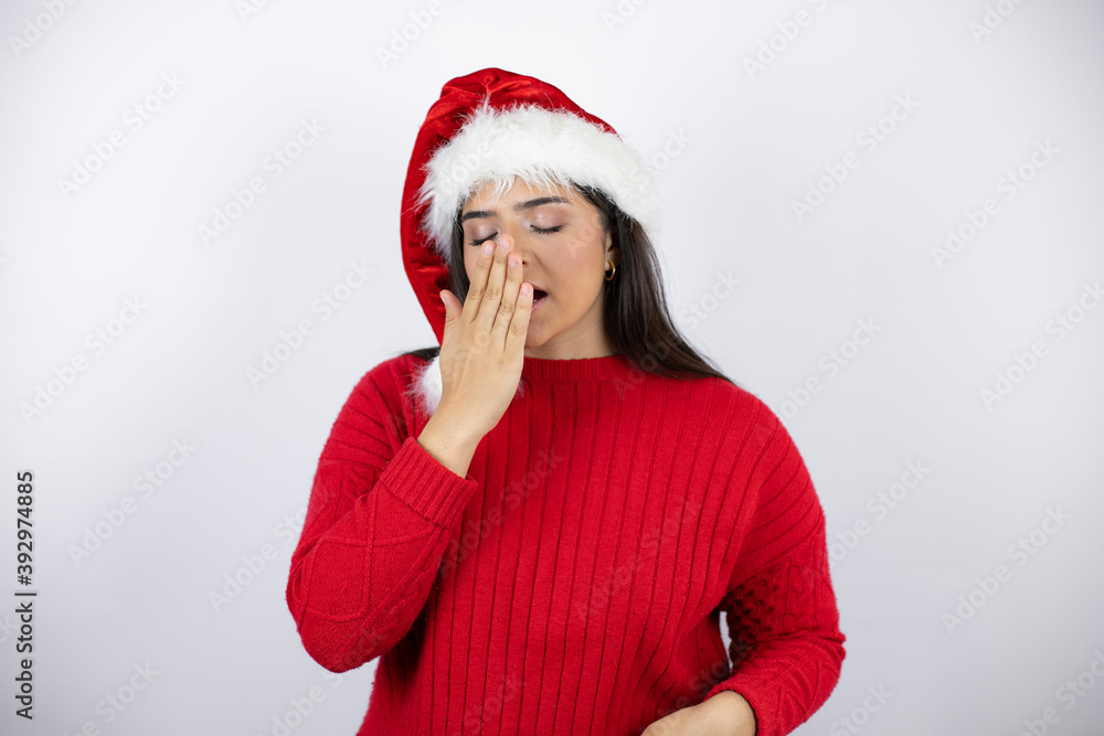Young beautiful woman wearing a Santa hat over white background bored yawning tired covering mouth with hand. Restless and sleepiness. Young beautiful woman wearing a Santa hat over white background bored yawning tired covering mouth with hand. Restless and sleepiness.