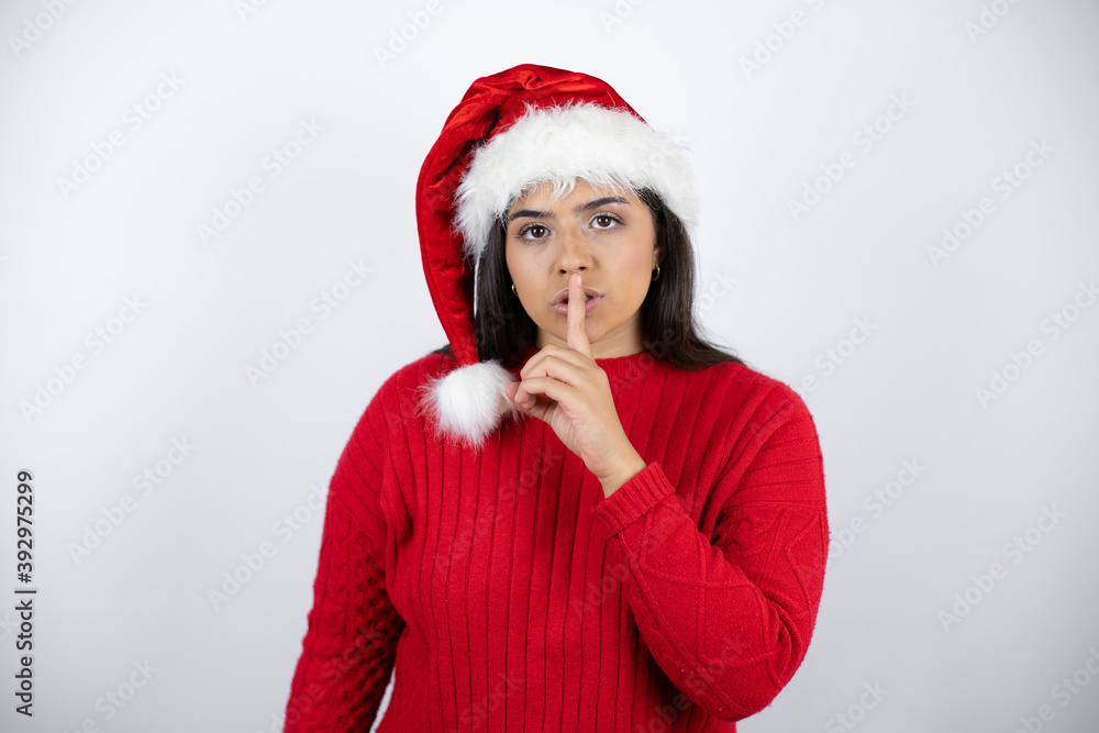 Young beautiful woman wearing a Santa hat over white background asking to be quiet with finger on lips. Silence and secret concept. Young beautiful woman wearing a Santa hat over white background asking to be quiet with finger on lips. Silence and secret concept.
