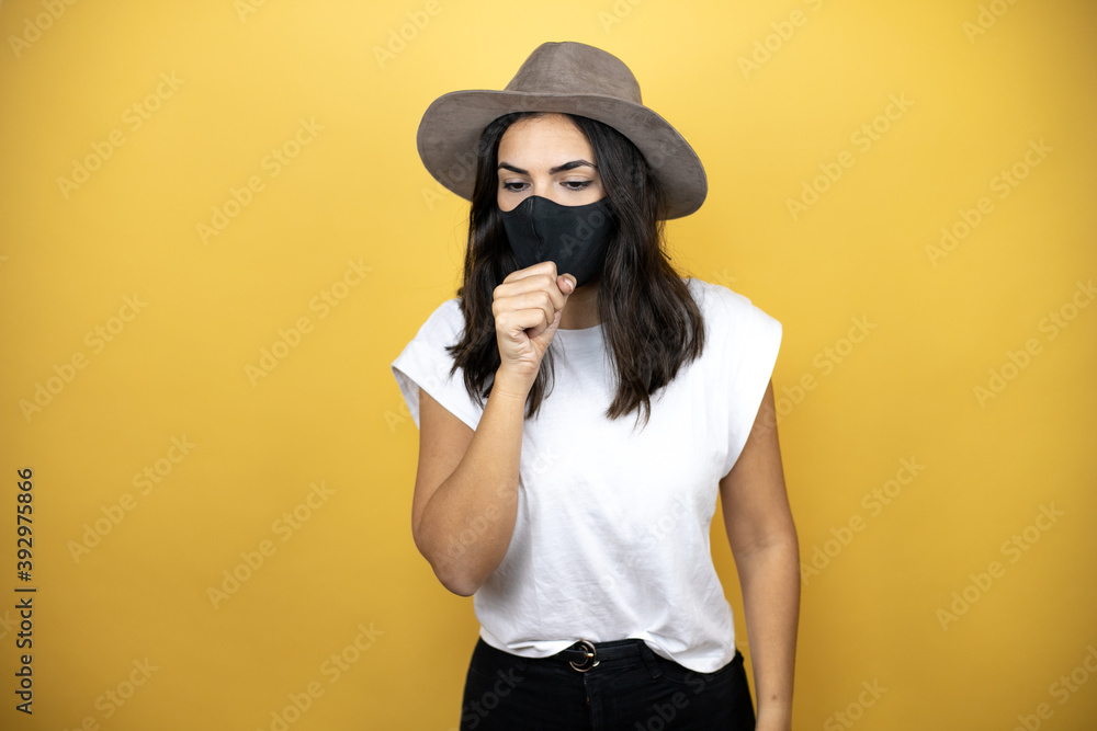 Beautiful woman wearing casual white t-shirt and a hat standing over yellow background with medical mask coughing Beautiful woman wearing casual white t-shirt and a hat standing over yellow background with medical mask coughing