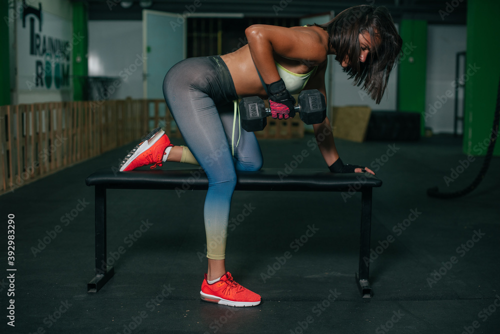 Fit young girl with light brown hair wearing black leggins, pink top and sneakers and doing squats with barbell, dark gym at background, cross training workout, portrait. Fit young girl with light brown hair wearing black leggins, pink top and sneakers and doing squats with barbell, dark gym at background, cross training workout, portrait.
