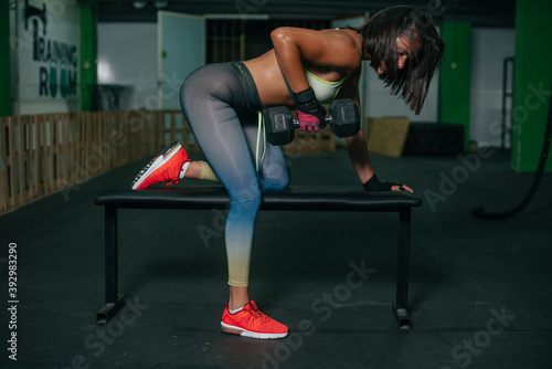 Fit young girl with light brown hair wearing black leggins, pink top and sneakers and doing squats with barbell, dark gym at background, cross training workout, portrait. © Bogdan